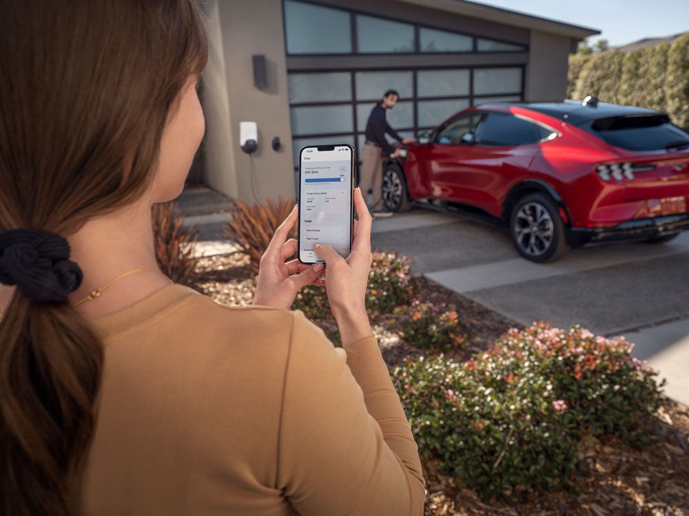 A woman on her phone with a man charging his Mustang Mach-E in the driveway